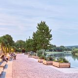 Flussufer mit von Bänken und Pflanzen umsäumter Uferpromenade mit Menschen auf Bänken sitzend